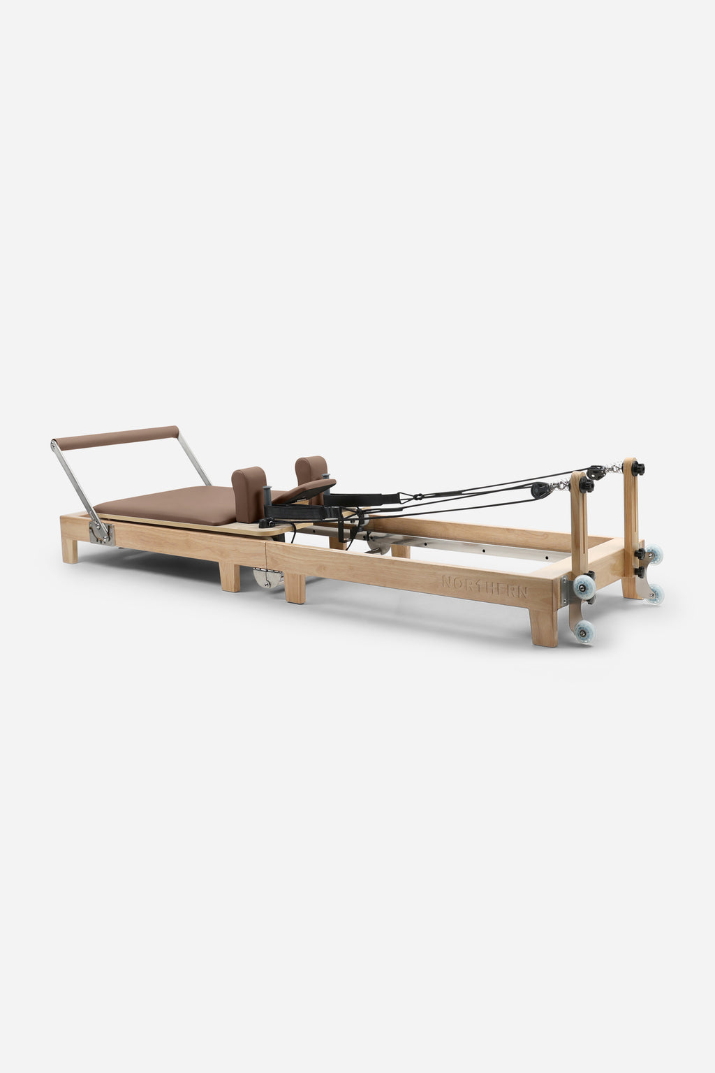 A Pilates reformer machine with a natural wood frame, brown cushioned bed and handles, and black springs, photographed on a clean light gray studio background.